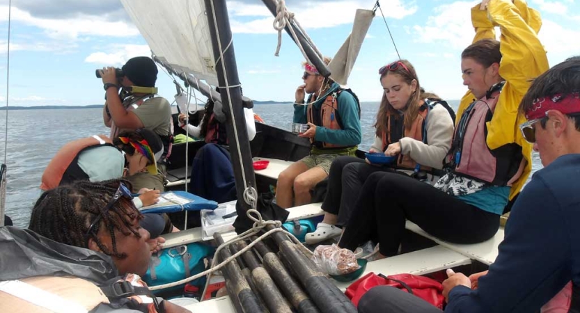 A group of people wearing life jackets sit on a sailboat. 